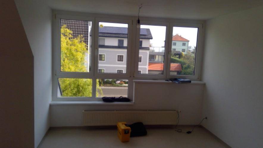 Heller Raum mit großen Fenstern und Blick auf die umliegenden Gebäude und Bäume.