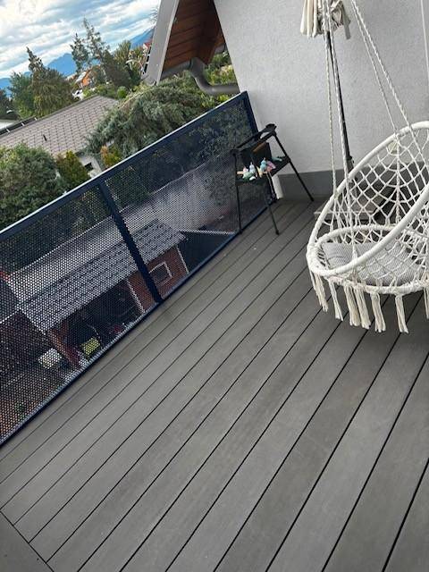 Moderner Balkon mit grauen Dielen, Hängesessel und weitem Blick über Dächer und Berge.