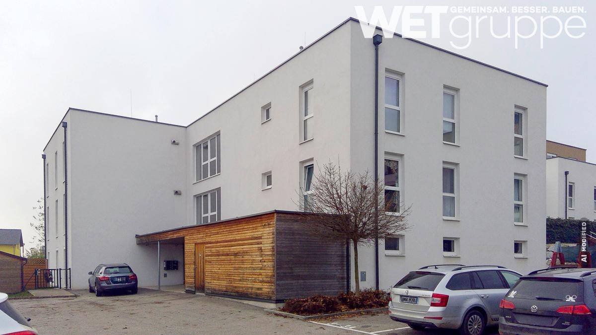 Modernes Mehrfamilienhaus mit weißer Fassade, großen Fenstern und Parkplätzen im Außenbereich.