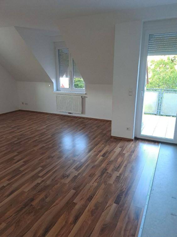 Geräumiges Zimmer mit dunklem Laminatboden, Dachschräge und Zugang zu einem Balkon.