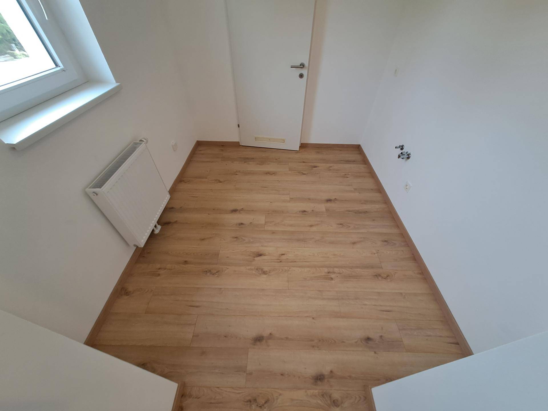 Leerer Raum mit hellem Laminatboden, einem Fenster und einem Heizkörper, bereit zur individuellen Gestaltung.