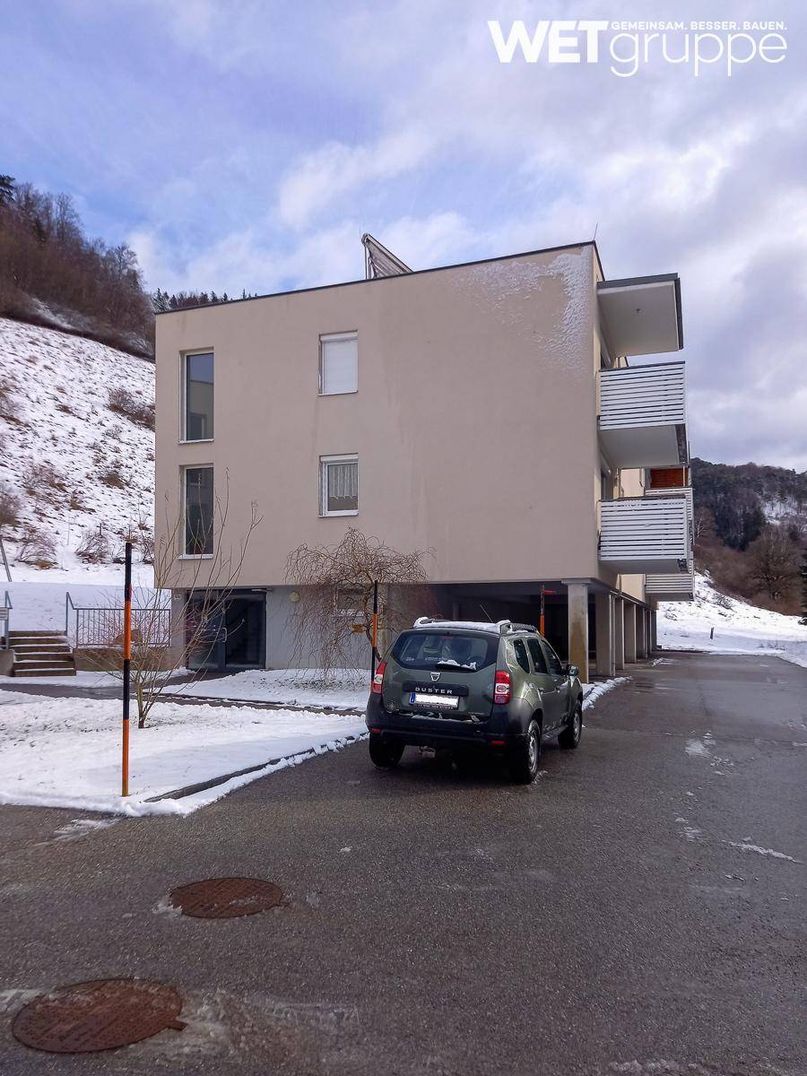 Modernes Wohngebäude mit Balkonen und einem geparkten Auto vor verschneiter Berglandschaft im Winter.