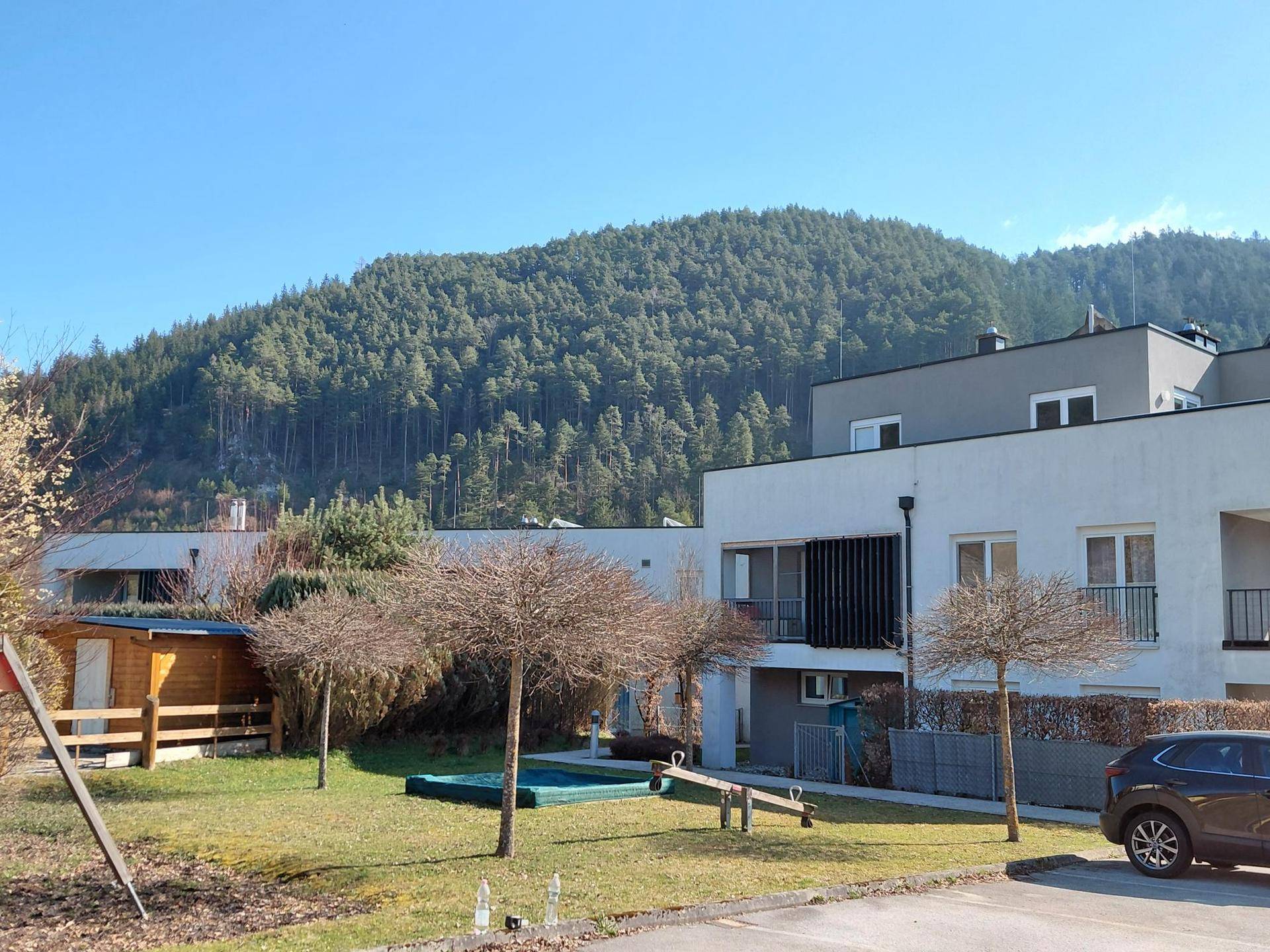 Moderne Wohnanlage mit Spielplatz und Blick auf bewaldete Berge unter blauem Himmel.