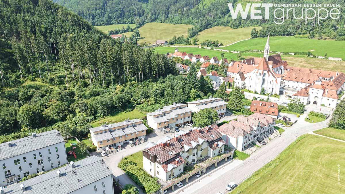 Luftaufnahme eines Dorfes mit Wohngebäuden, umgeben von grünen Hügeln und Wäldern, mit einer historischen Kirche.
