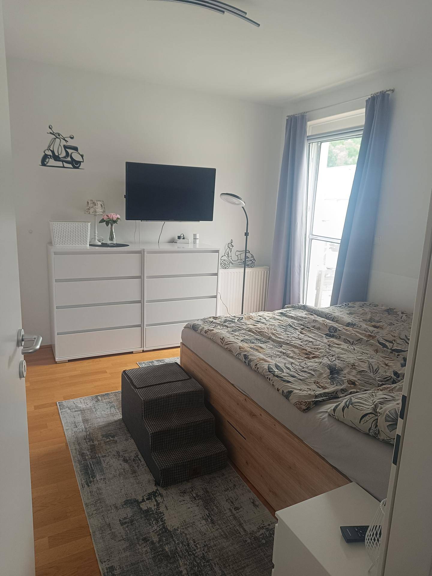 Gemütliches Schlafzimmer mit Doppelbett, weißen Kommoden und Fenster mit blauem Vorhang.