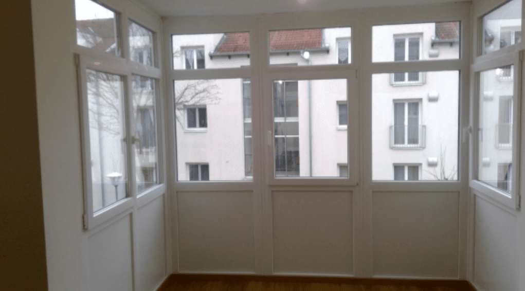 Helle Loggia mit vielen Fenstern, Holzboden und Blick auf umliegende Gebäude.