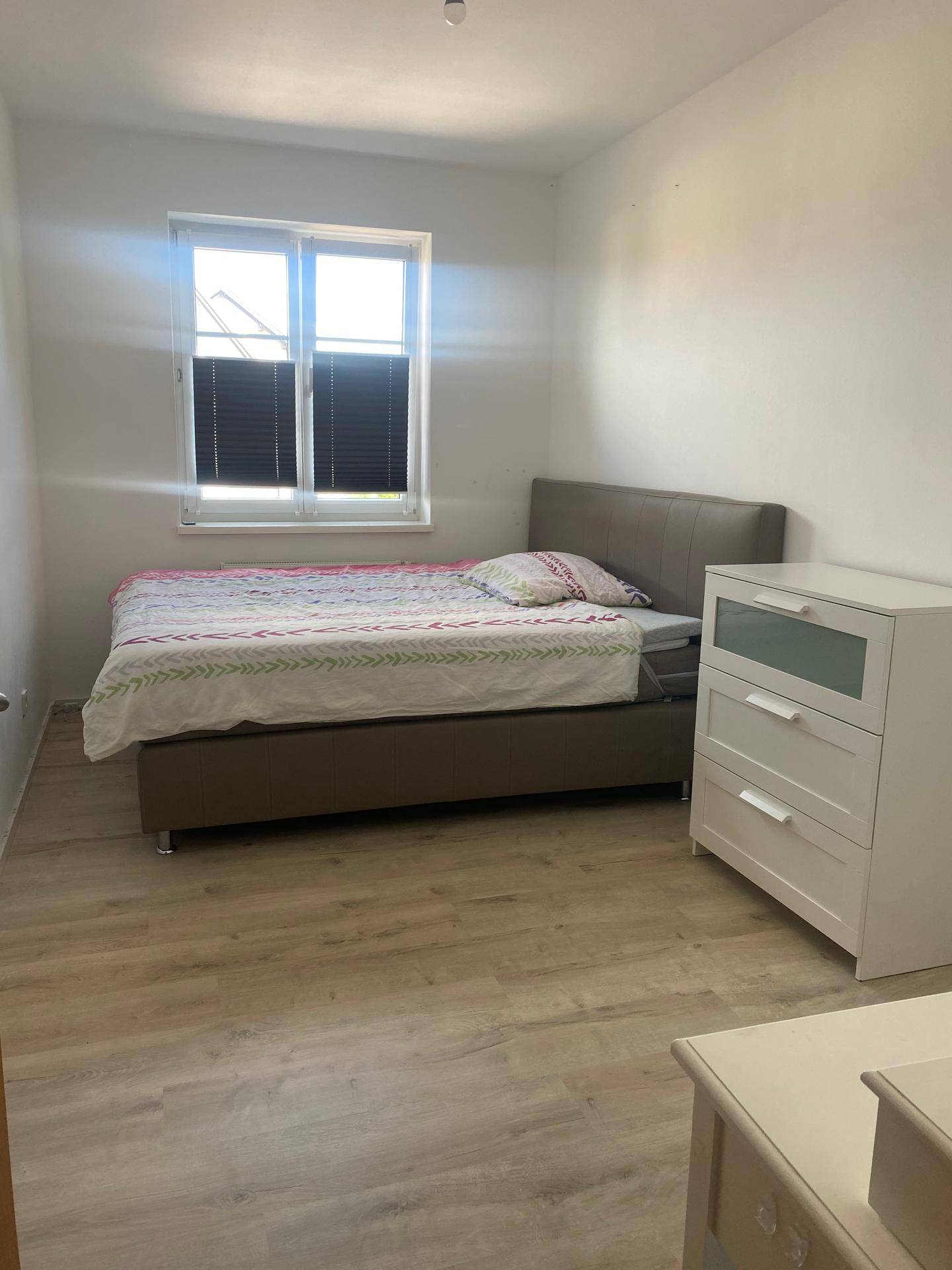 Helles Schlafzimmer mit einem bequemen Bett, einem weißen Nachttisch und modernem Laminatboden.