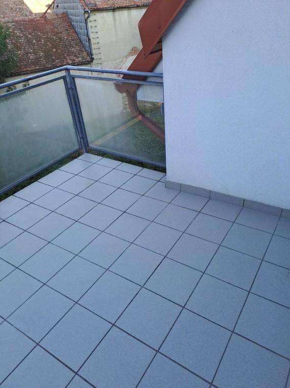 Balkon mit grauen Fliesen und Glasgeländer, bietet Blick auf die umliegenden Dächer.