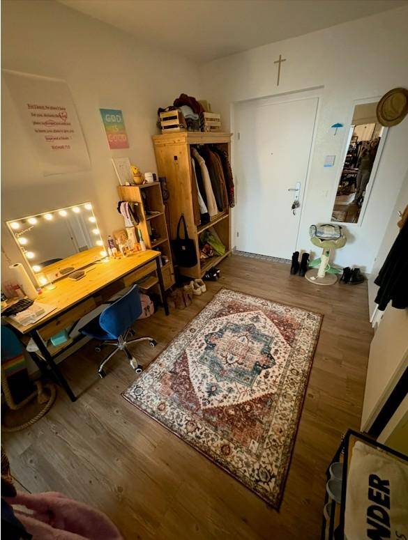 Helles Zimmer mit Schminktisch, offenem Kleiderschrank und gemustertem Teppich.