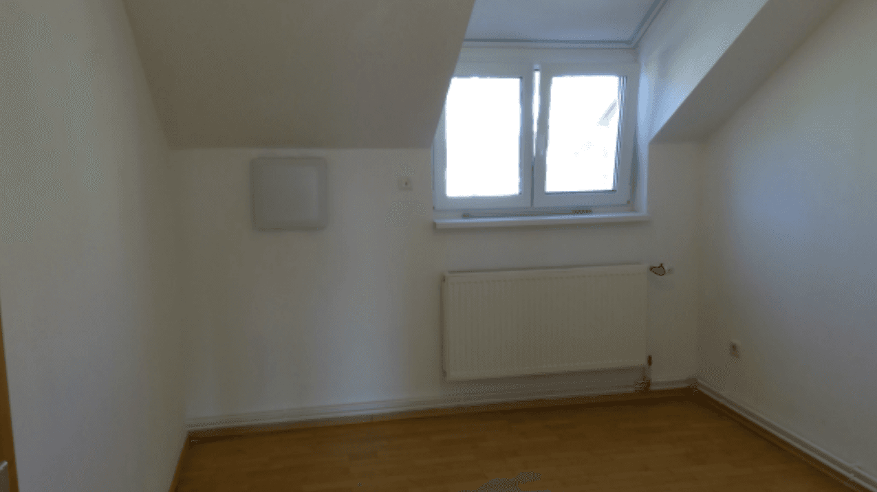 Helles Zimmer im Dachgeschoss mit Schrägdecken, Laminatboden und Heizkörper unter dem Fenster.