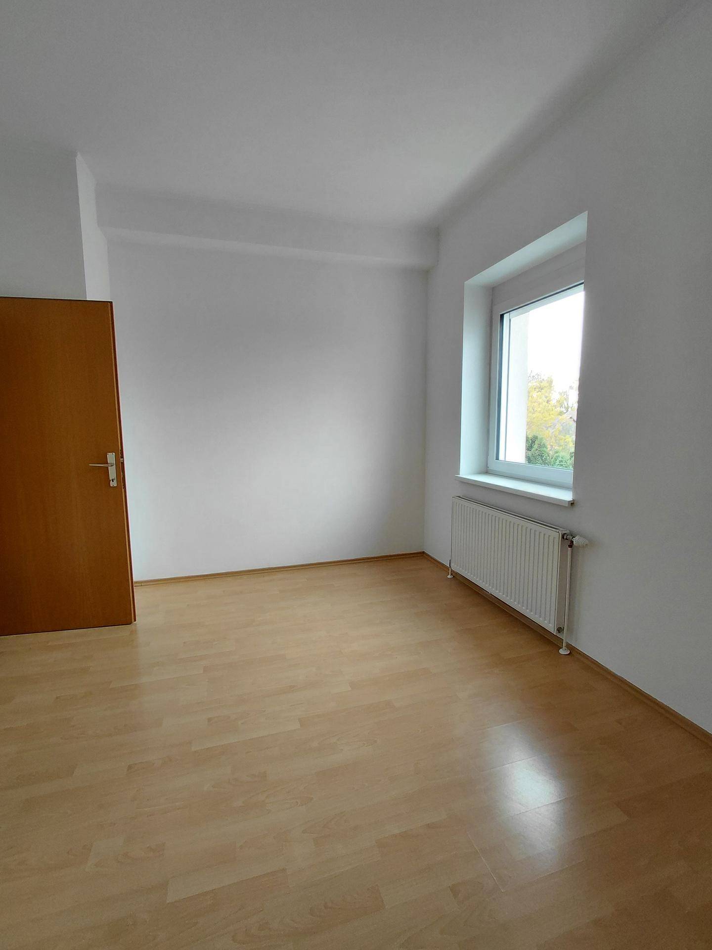 Helles Zimmer mit hellem Laminatboden, weißen Wänden und einem Fenster mit Heizkörper.
