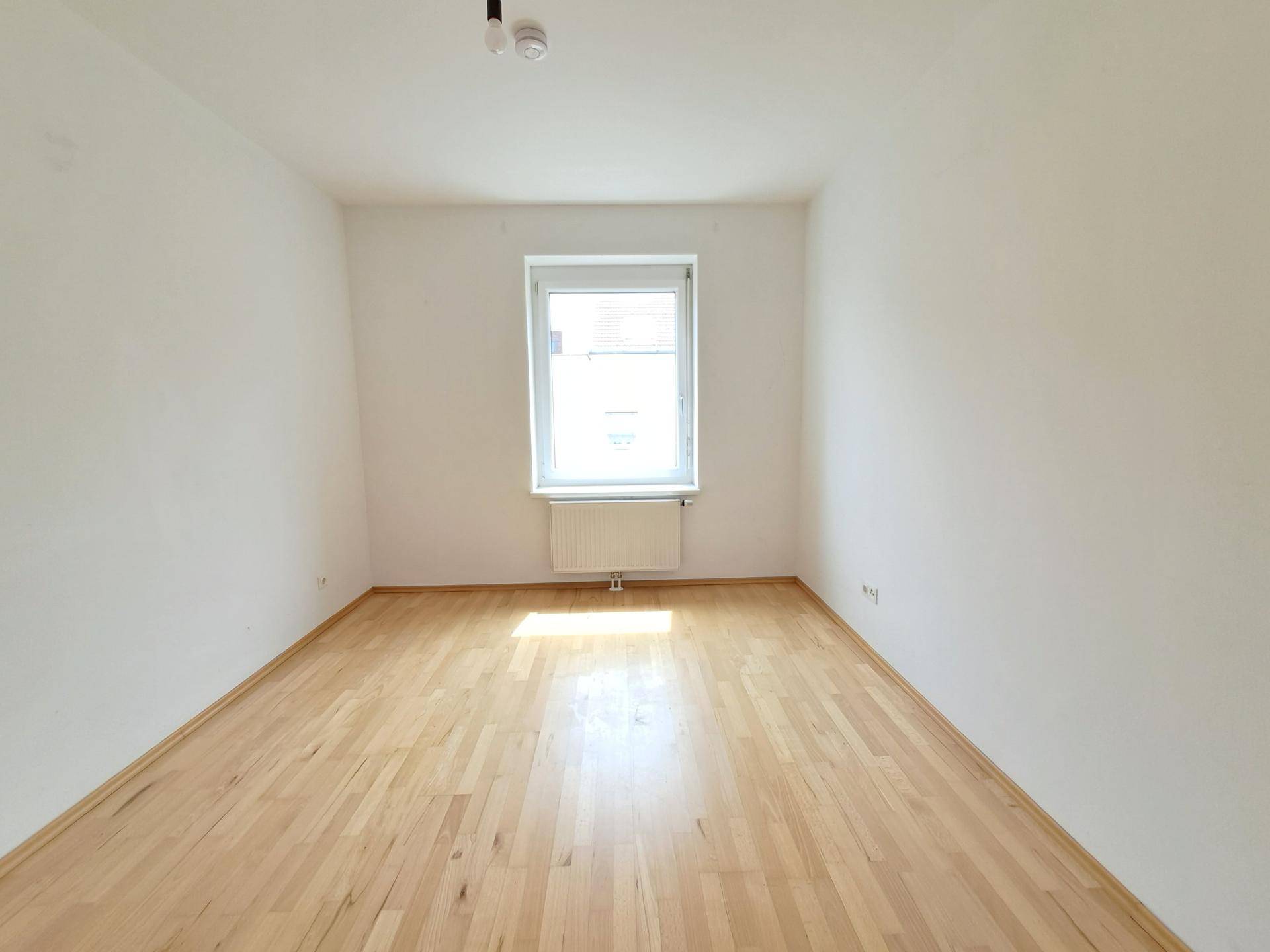 Leeres Zimmer mit hellem Holzboden, einem Fenster und Heizkörper, bereit zur Einrichtung.