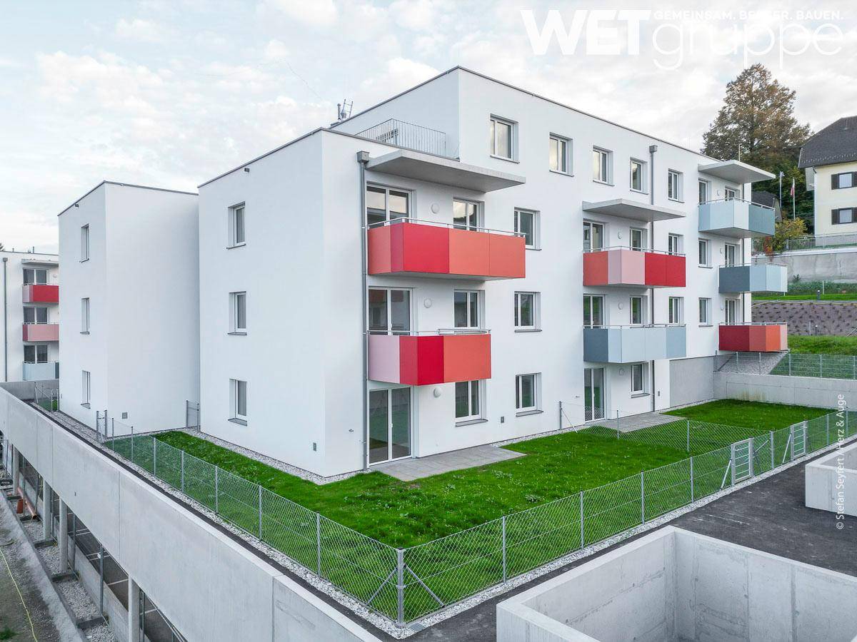 Ansicht eines modernen Wohngebäudes mit farbigen Balkonen und einer umzäunten Rasenfläche.