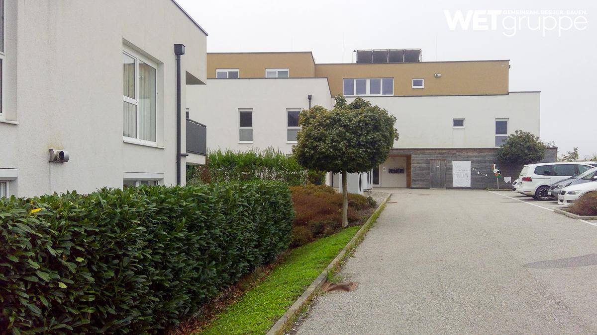 Seitenansicht eines modernen Gebäudes mit Parkplatz, Hecke und einem kleinen Baum im Vordergrund.