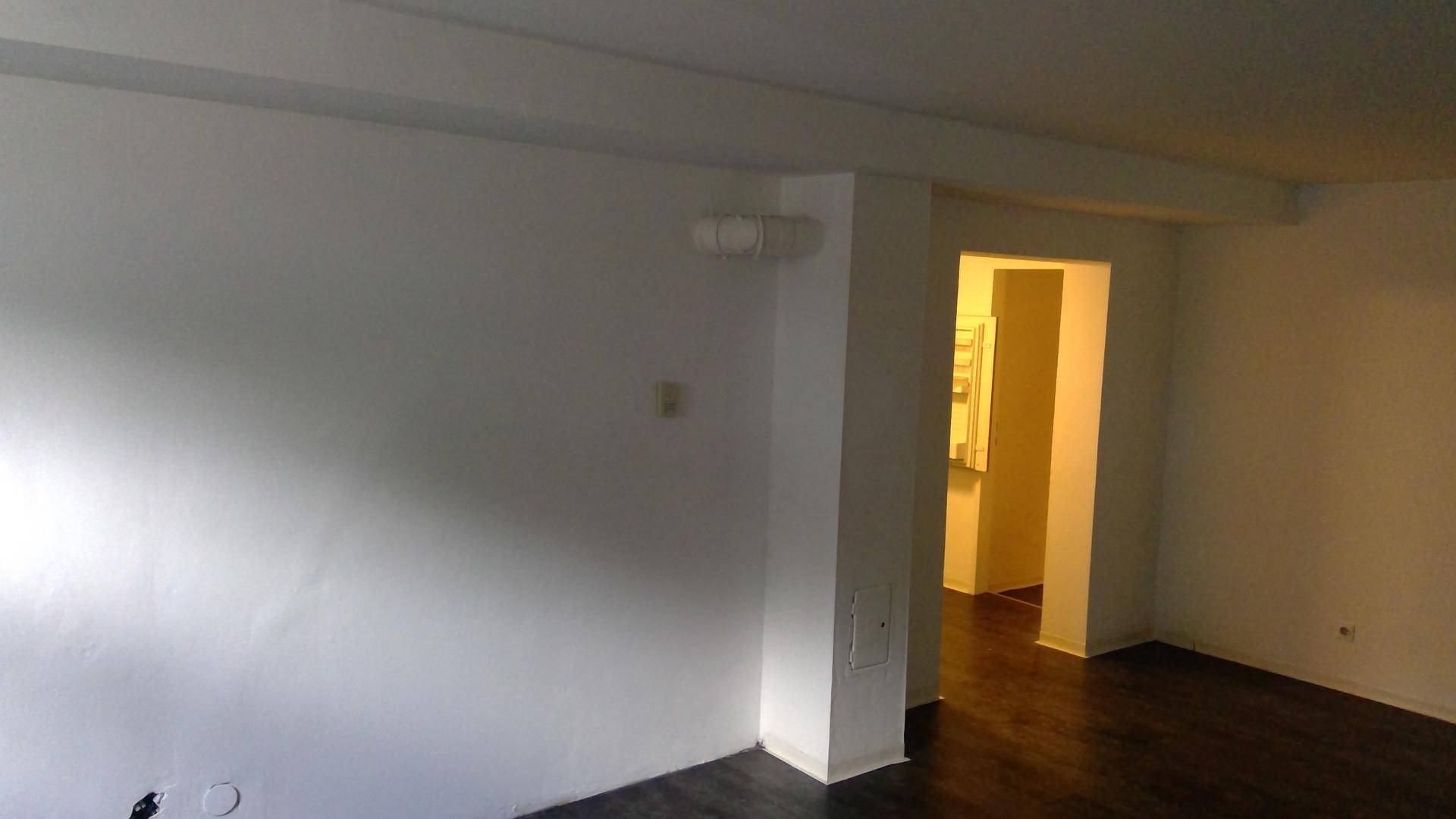 Wohnbereich mit dunklem Laminatboden, weißen Wänden und einem Durchgang zu einem weiteren Raum.