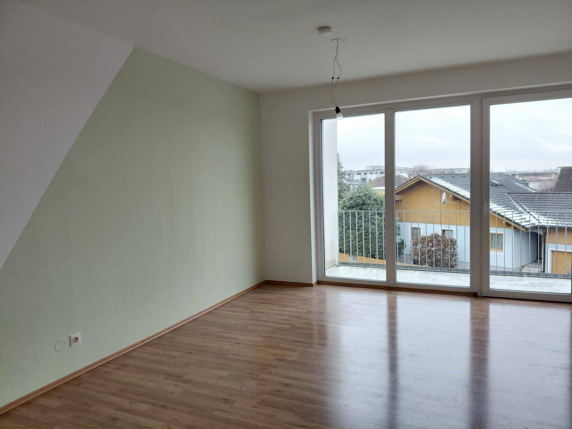 Geräumiger Wohnraum mit Laminatboden, großem Fenster und Zugang zum Balkon, mit Dachschräge.