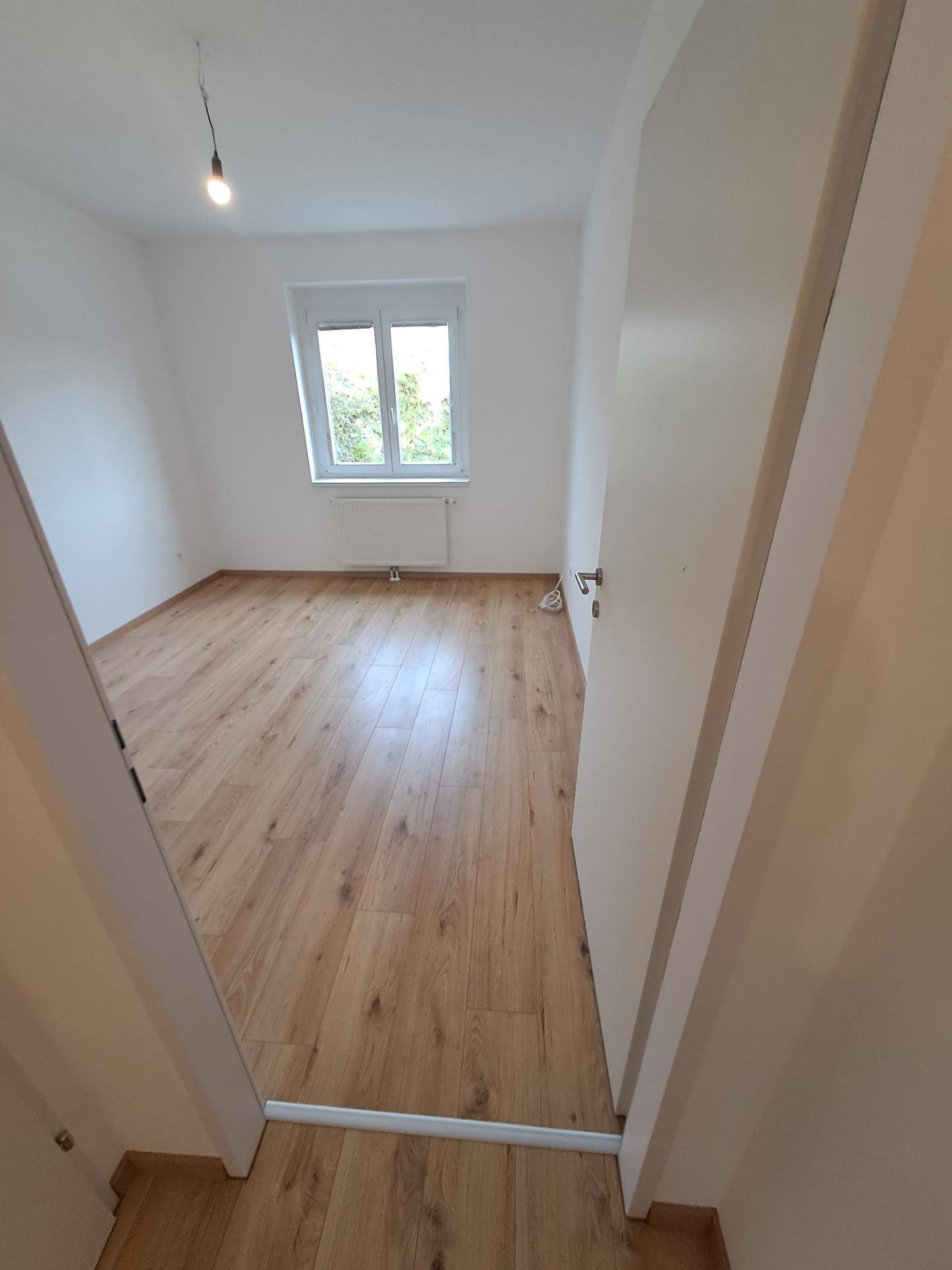 Schlafzimmer mit hellem Laminatboden, einem Fenster mit Blick ins Grüne und einem Heizkörper.