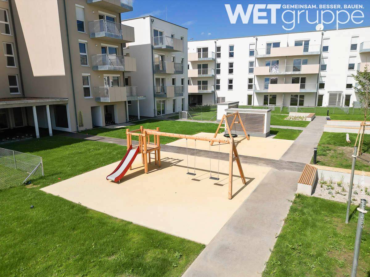 Detailaufnahme des Spielplatzes im Innenhof mit Schaukeln und Rutsche, umgeben von Wohngebäuden.