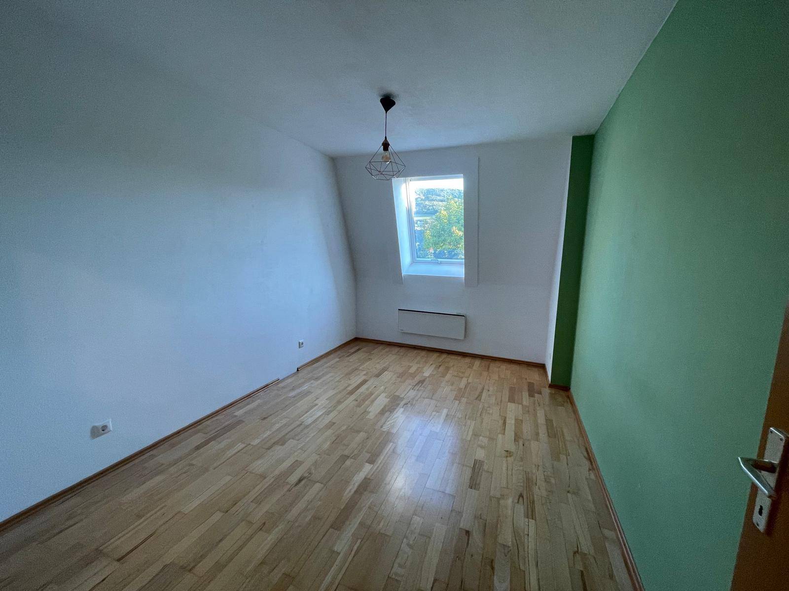 Helles Zimmer mit Holzboden, grüner Akzentwand und Fenster in der Dachschräge.