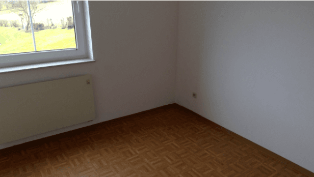 Leeres Zimmer mit hellem Parkettboden, weißen Wänden, Fenster und Heizkörper, mit Blick ins Grüne.