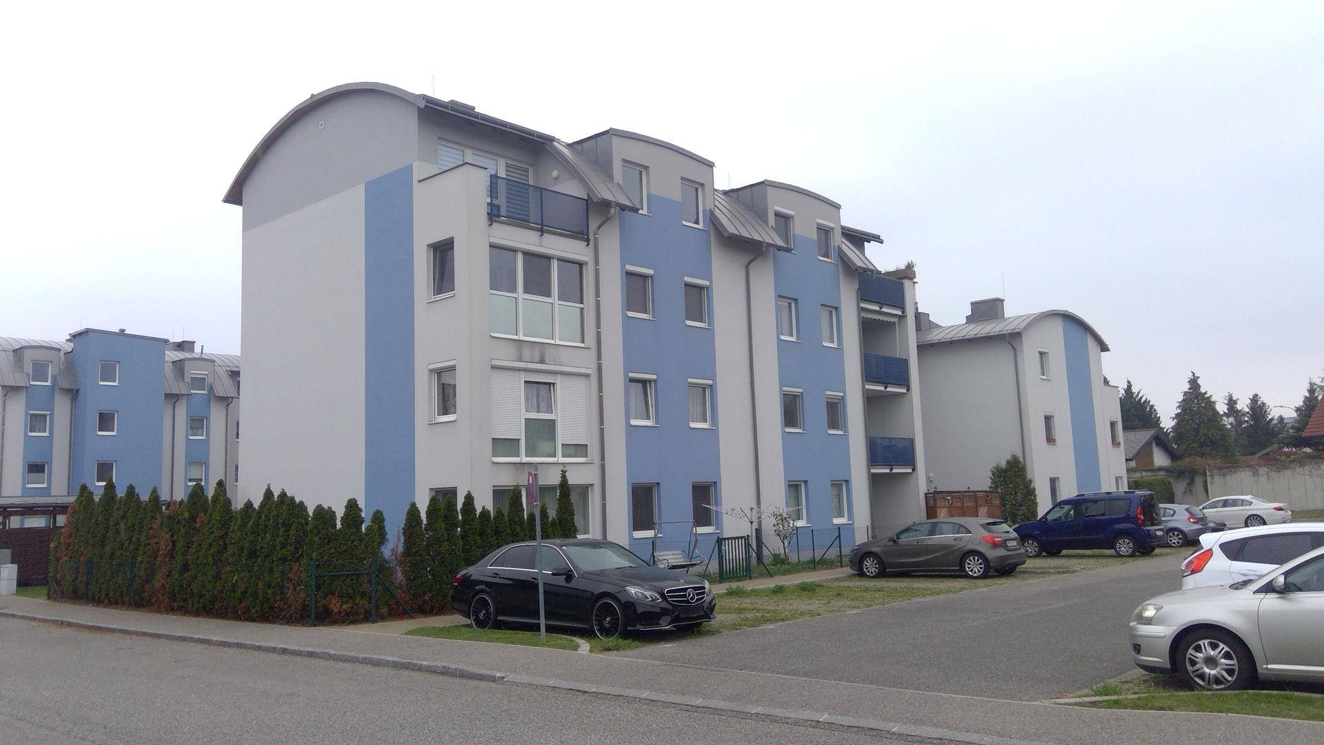 Wohngebäude mit blauer und grauer Fassade, umgeben von Grünflächen und Parkplätzen.