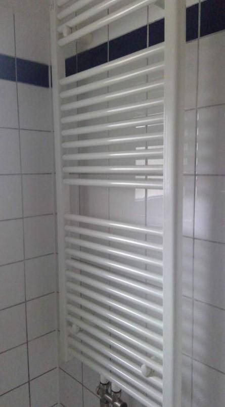 Weißer Handtuchheizkörper in einem Badezimmer mit weißen Fliesen und blauer Akzentlinie an der Wand.