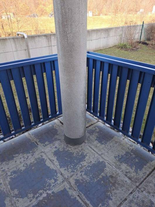 Kleiner Balkon mit blauer Brüstung und abgenutztem Bodenbelag, umgeben von Grünflächen.