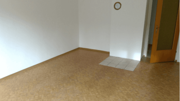 Leerer Wohnraum mit hellem, gemustertem Bodenbelag und einem kleinen Fliesenbereich am Eingang.