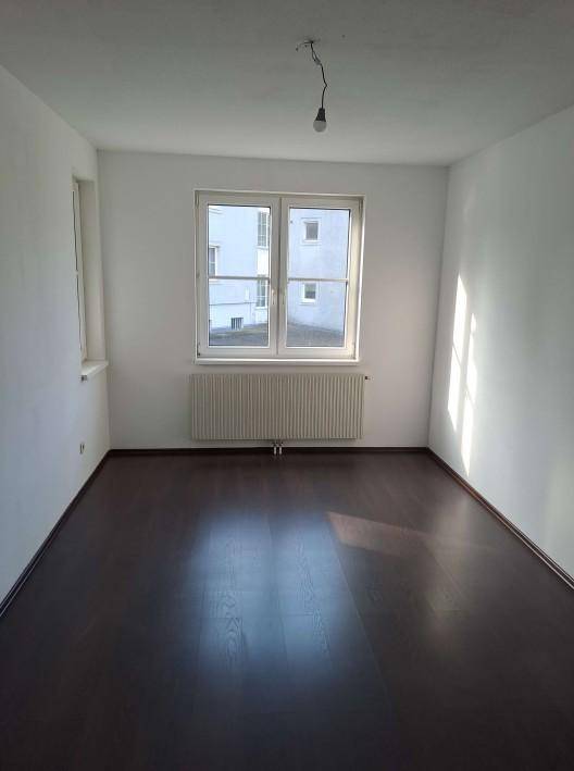 Leerer Raum mit dunklem Laminatboden, weißen Wänden und einem Fenster mit Heizkörper darunter.