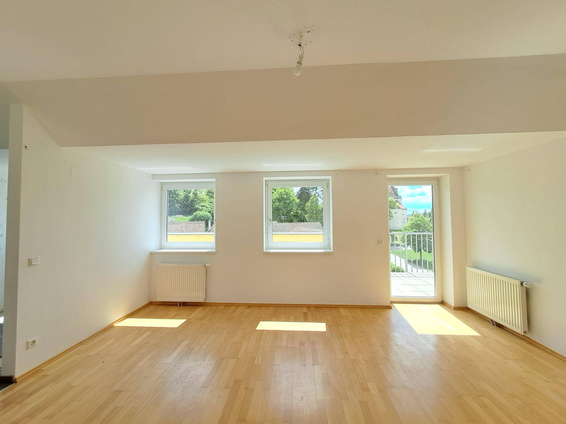 Geräumiges Wohnzimmer mit Holzboden, großen Fenstern und Zugang zu einem Balkon.