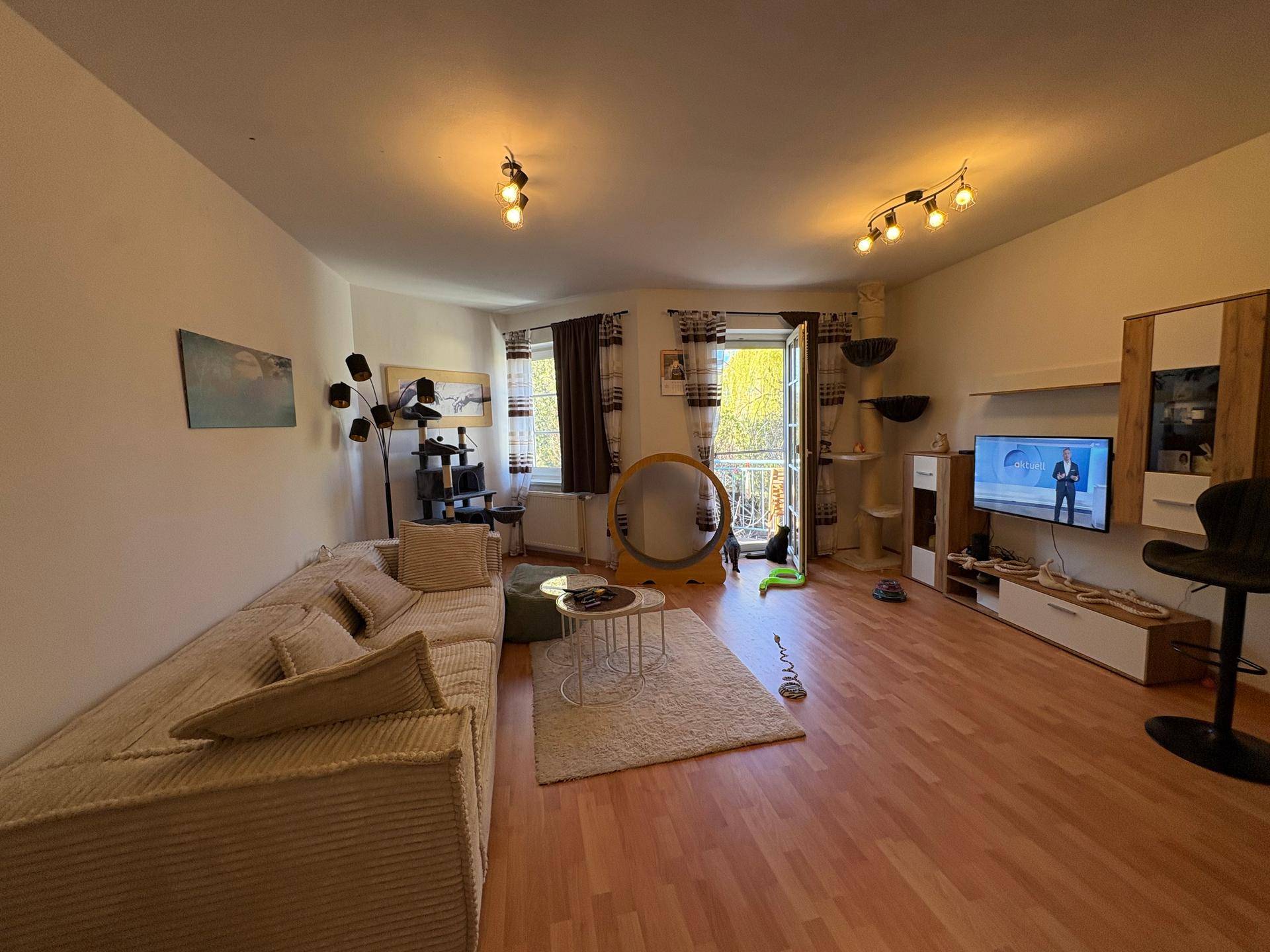Helles Wohnzimmer mit beigem Sofa, Laminatboden und Zugang zum Balkon.