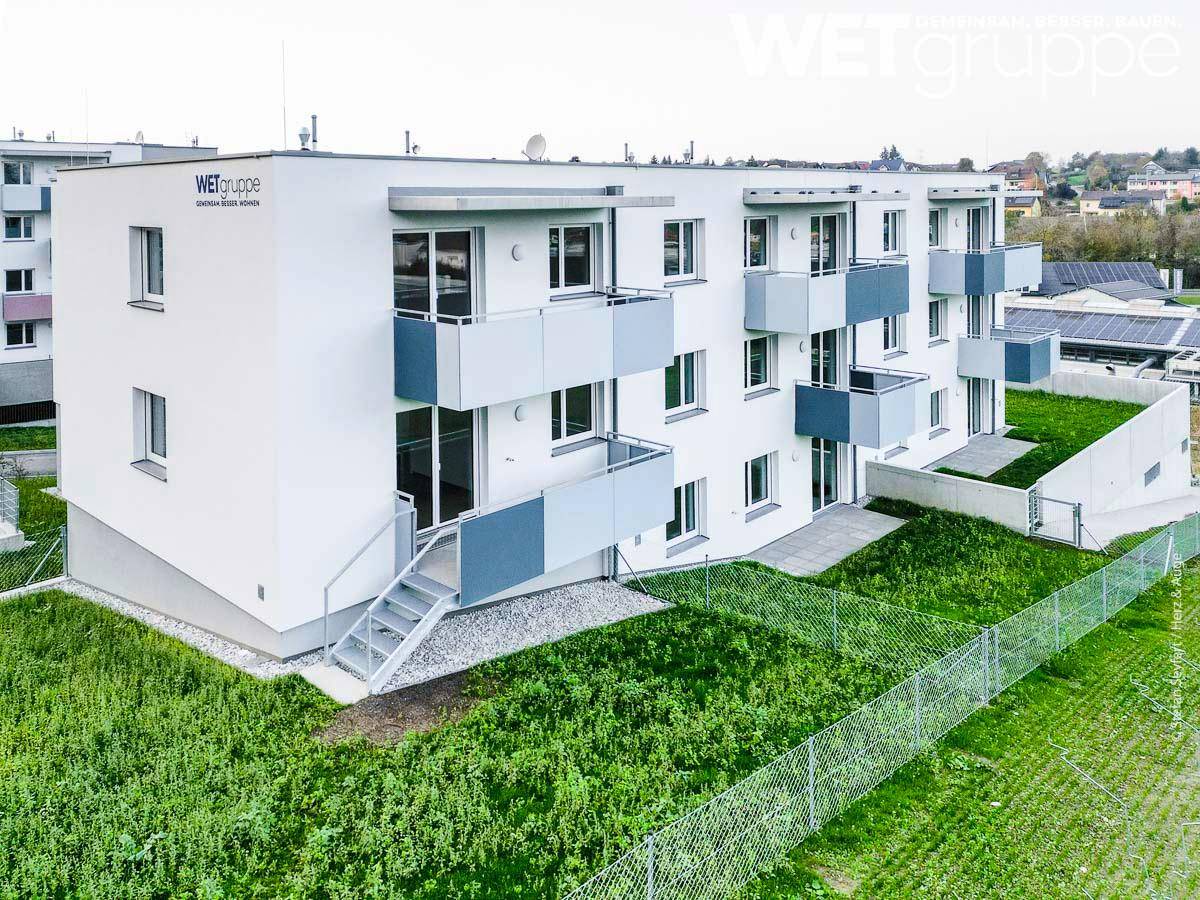 Modernes Wohnhaus mit schlichter weißer Fassade, grauen Balkonen und umliegenden Grünflächen.