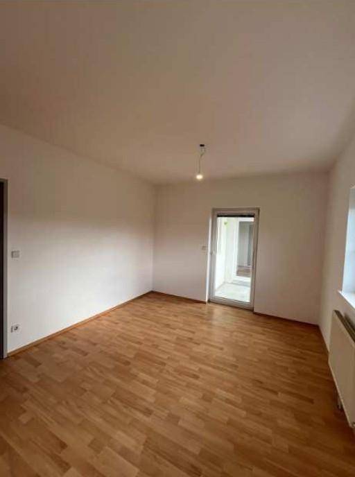 Leerer Raum mit hellem Holzfußboden, weißen Wänden und Zugang zu einem weiteren Bereich.