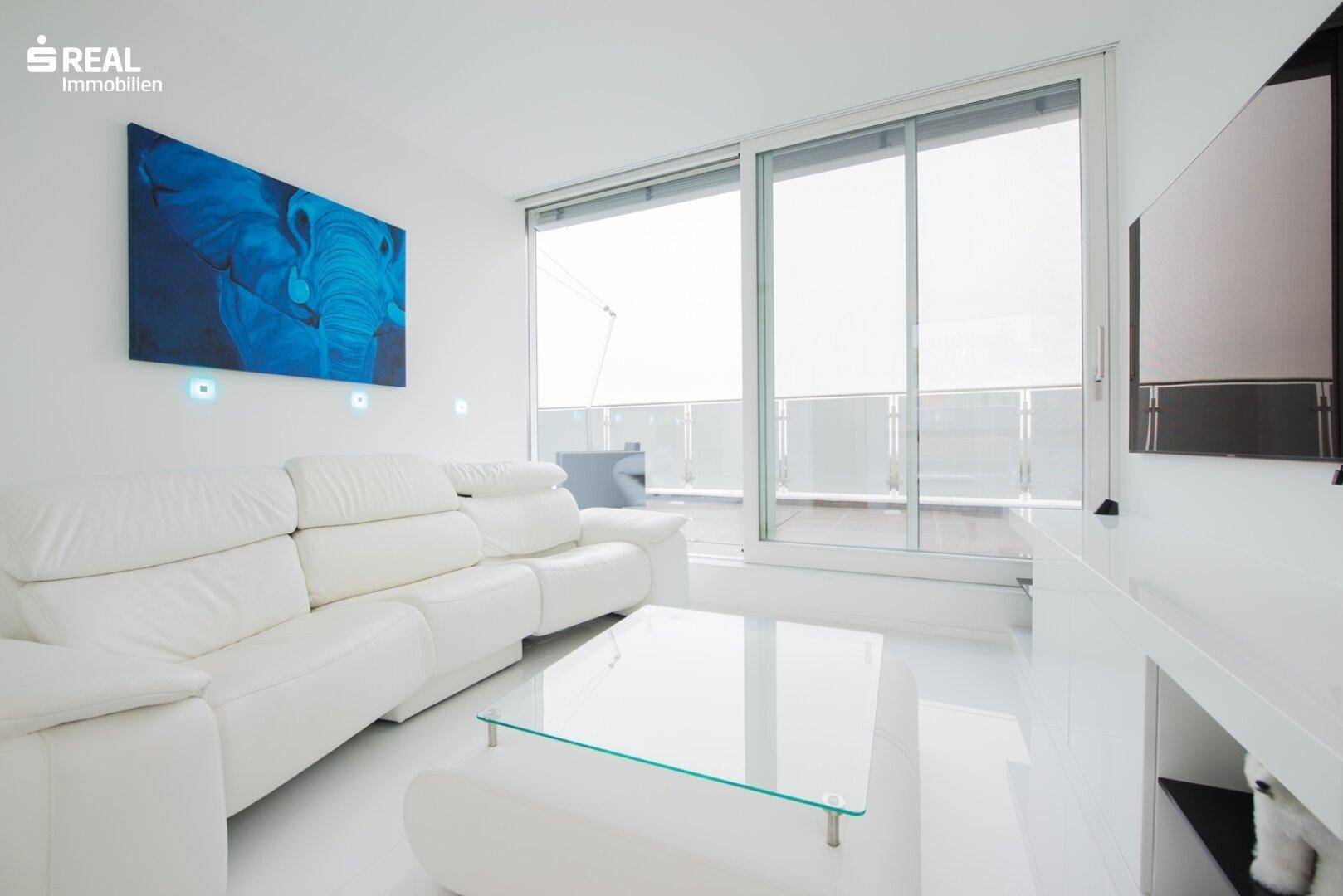 Helles Wohnzimmer mit weißem Ledersofa, Glastisch und Zugang zu einem Balkon mit Weitblick.