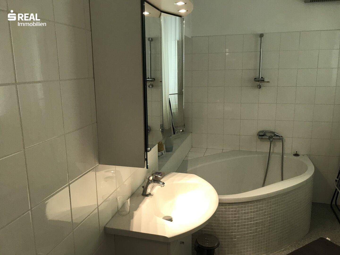 Badezimmer mit weißen Fliesen, einer Eckbadewanne und einem einzelnen Waschbecken.