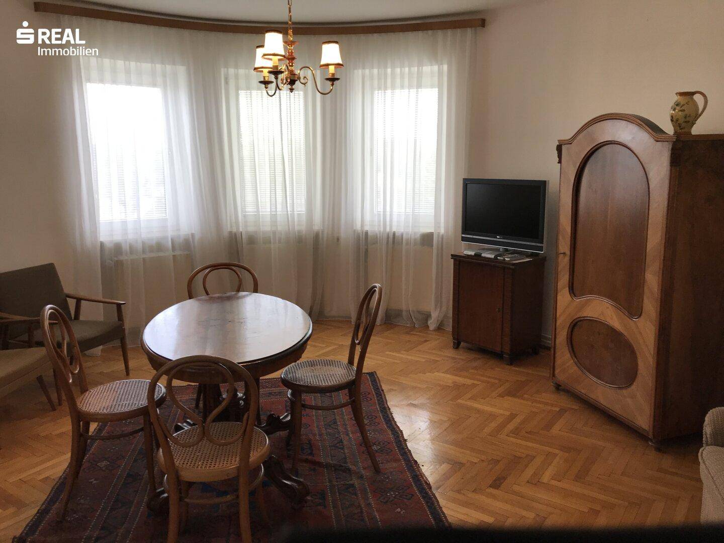 Wohnzimmer mit rundem Holztisch, traditionellem Schrank und Parkettboden.