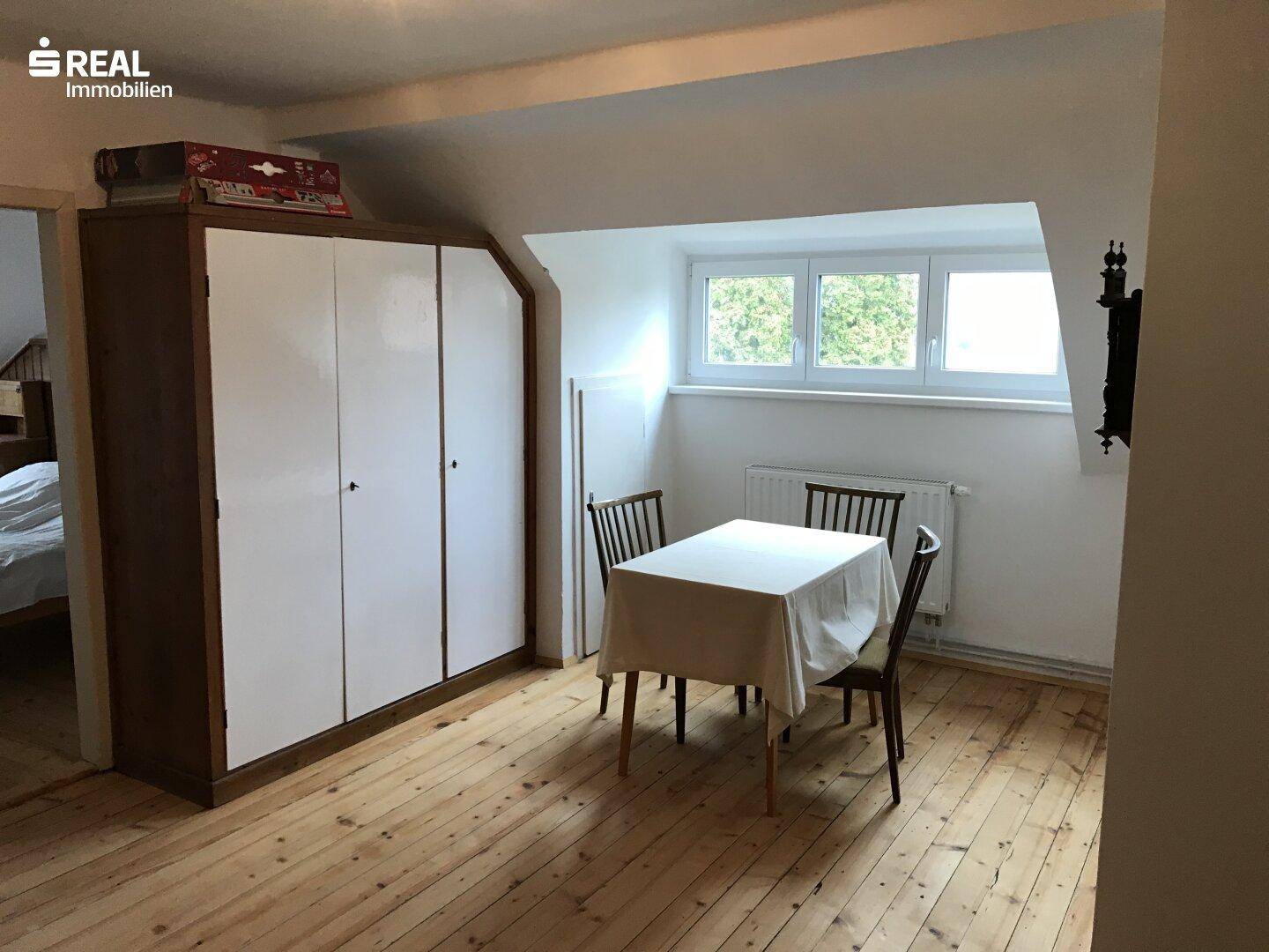 Dachzimmer mit Holzdielenboden, Schrank und kleinem Esstisch.