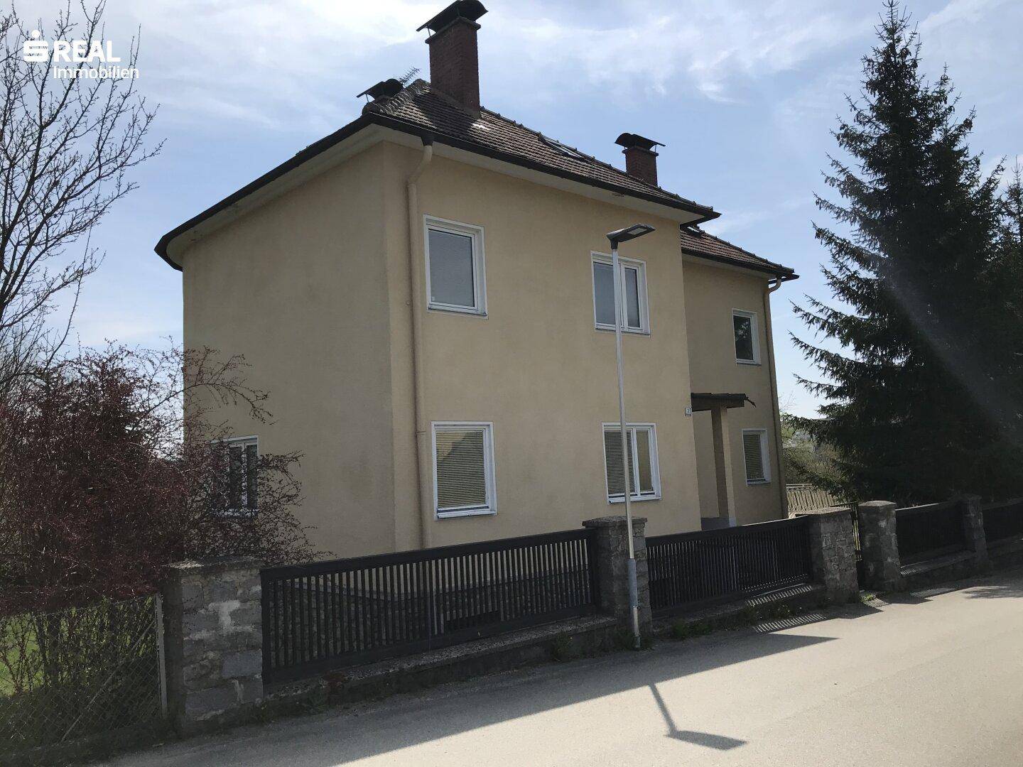 Vorderansicht des Hauses von der Straße aus, mit einem Zaun und Bäumen im Hintergrund.