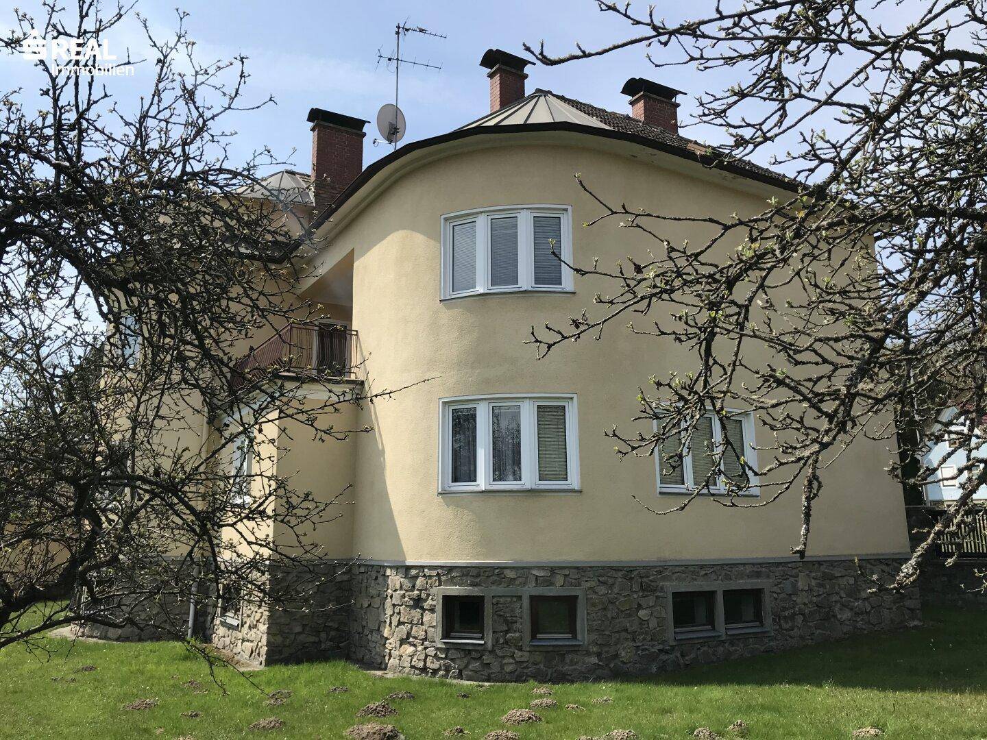 Historisches Haus mit gelber Fassade und markanten Schornsteinen umgeben von Bäumen.