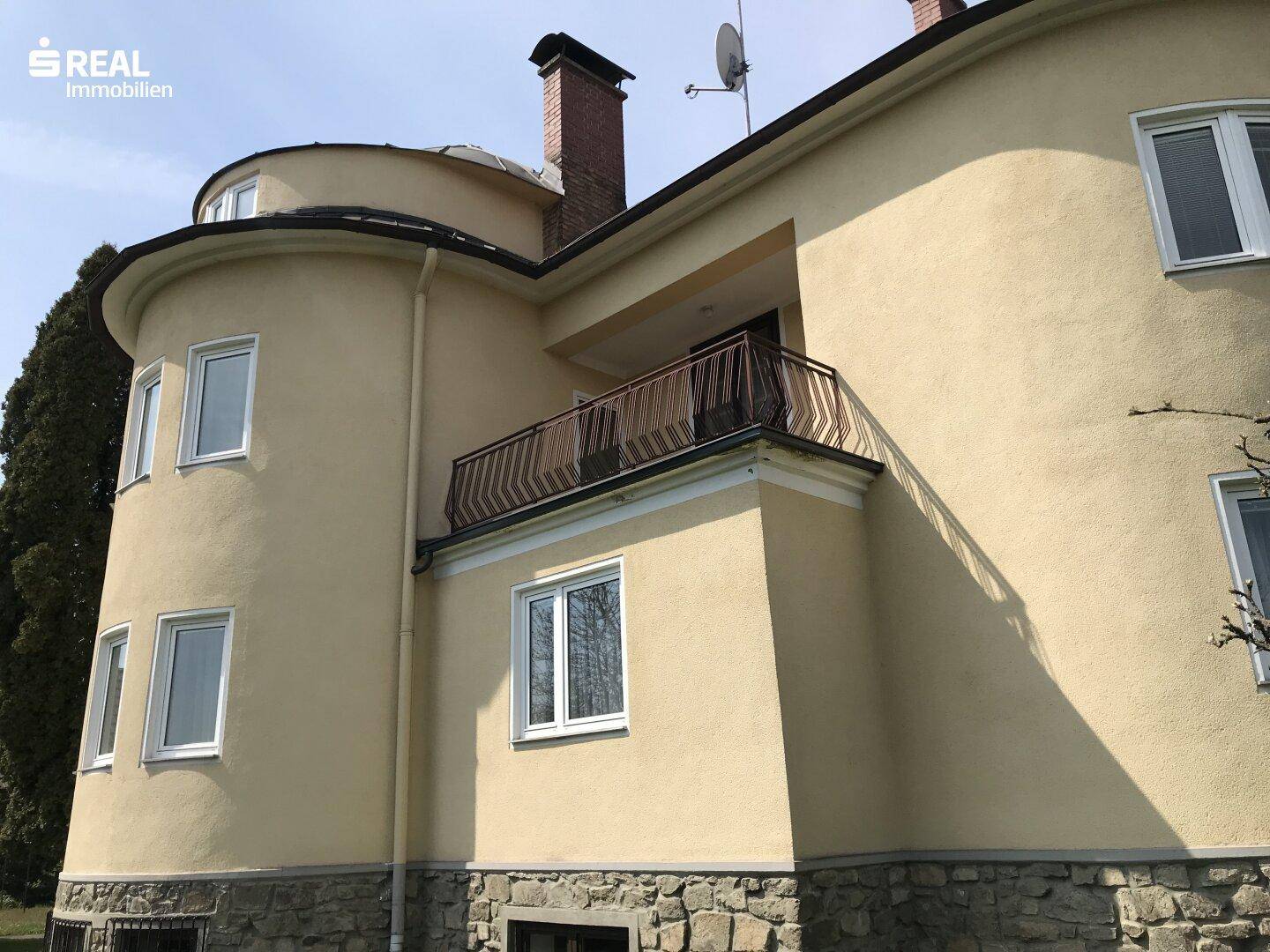 Detailansicht der gelben Hausfassade mit einem kleinen Balkon und schmiedeeisernem Geländer.