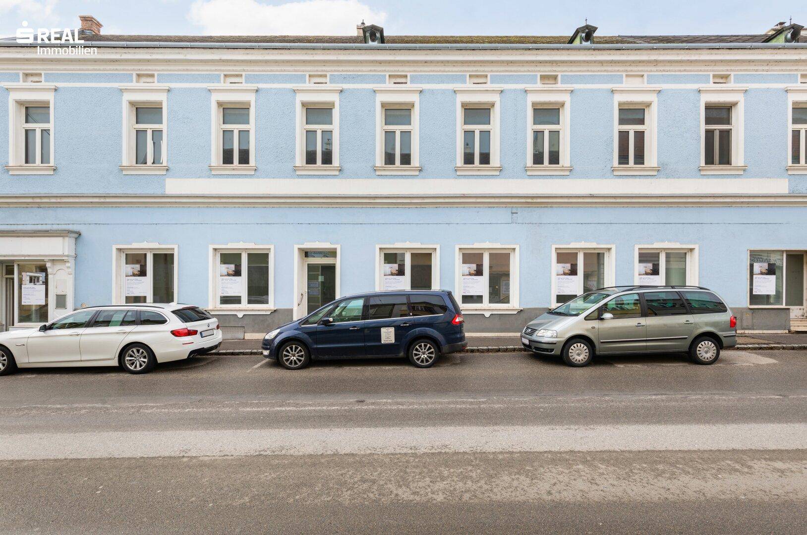 Historisches Gebäude mit hellblauer Fassade und vielen Fenstern entlang einer Straße mit parkenden Autos.