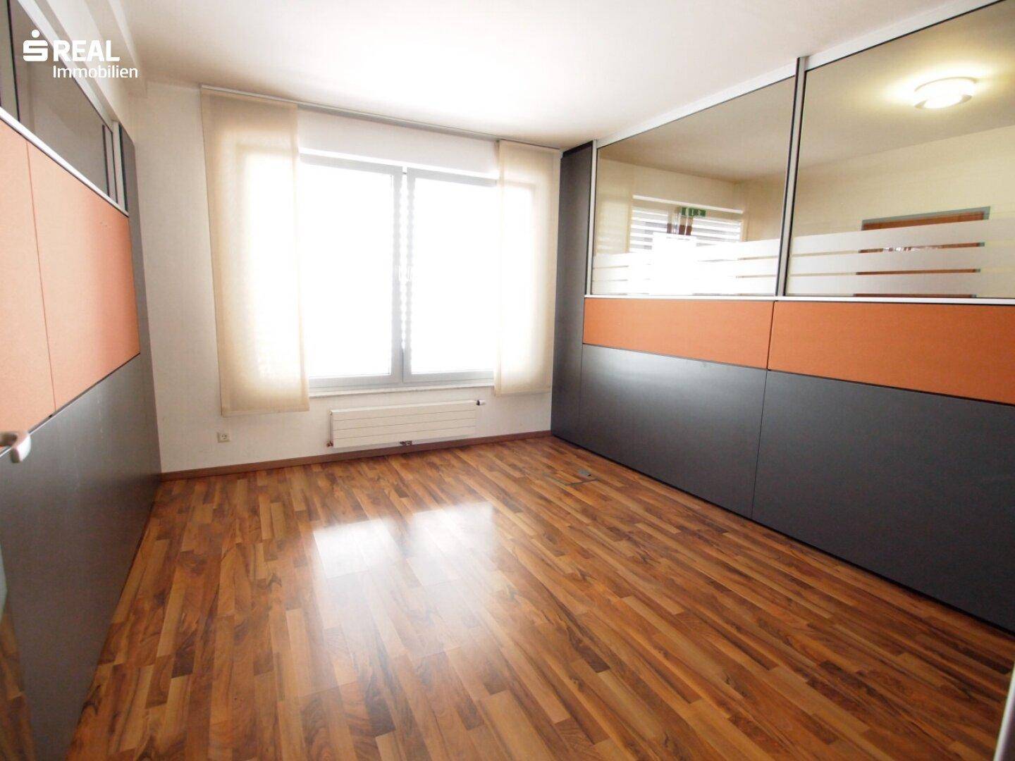 Heller Büroraum mit Holzboden, großem Fenster und modernen Trennwänden in Grau und Orange.
