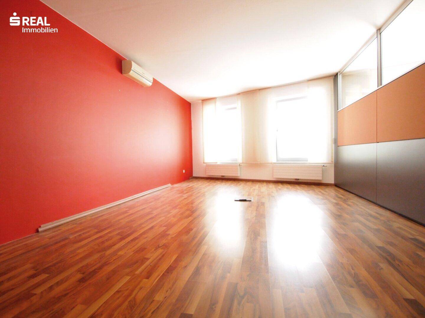 Geräumiger Bürobereich mit roter Akzentwand, Holzboden und mehreren Fenstern für viel Tageslicht.