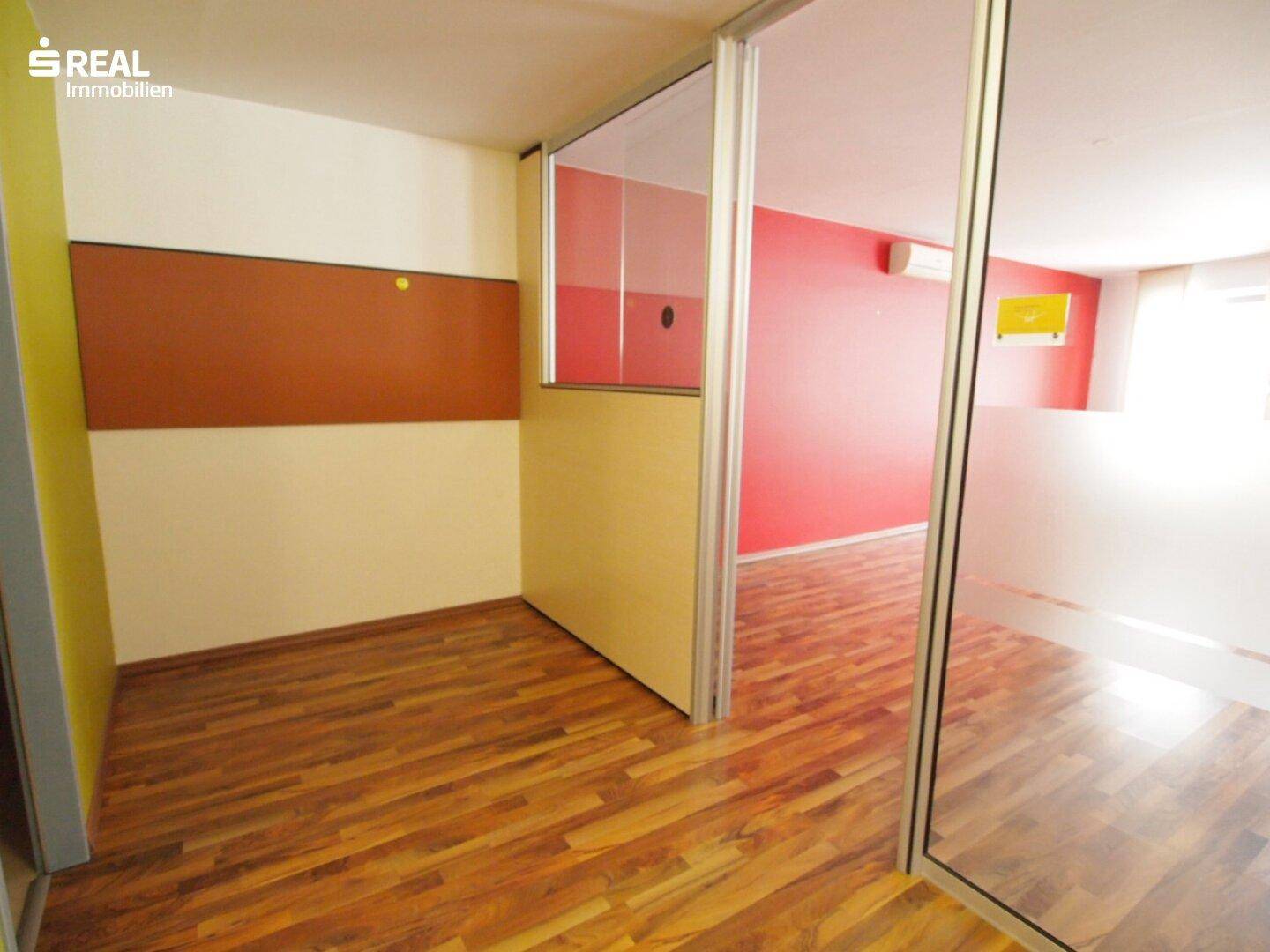 Büroraum mit Holzboden, gelber Akzentwand und Glastrennwänden, die den Blick in angrenzende Bereiche freigeben.