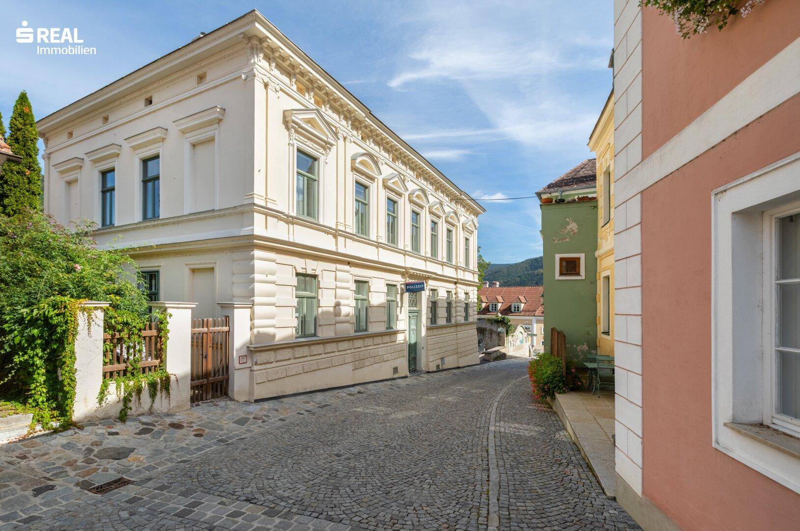 Elegantes historisches Gebäude an einer gepflasterten Straße mit grüner Bepflanzung und blauer Himmel.