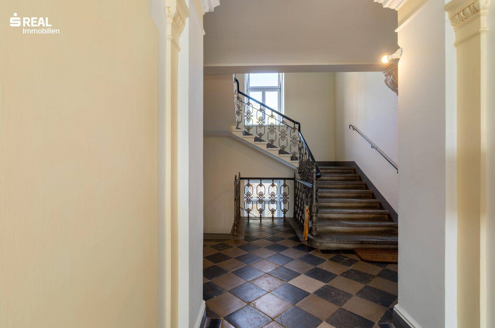 Historisches Treppenhaus mit kunstvollem Geländer, hohen Decken und Schachbrettmuster-Fliesenboden.