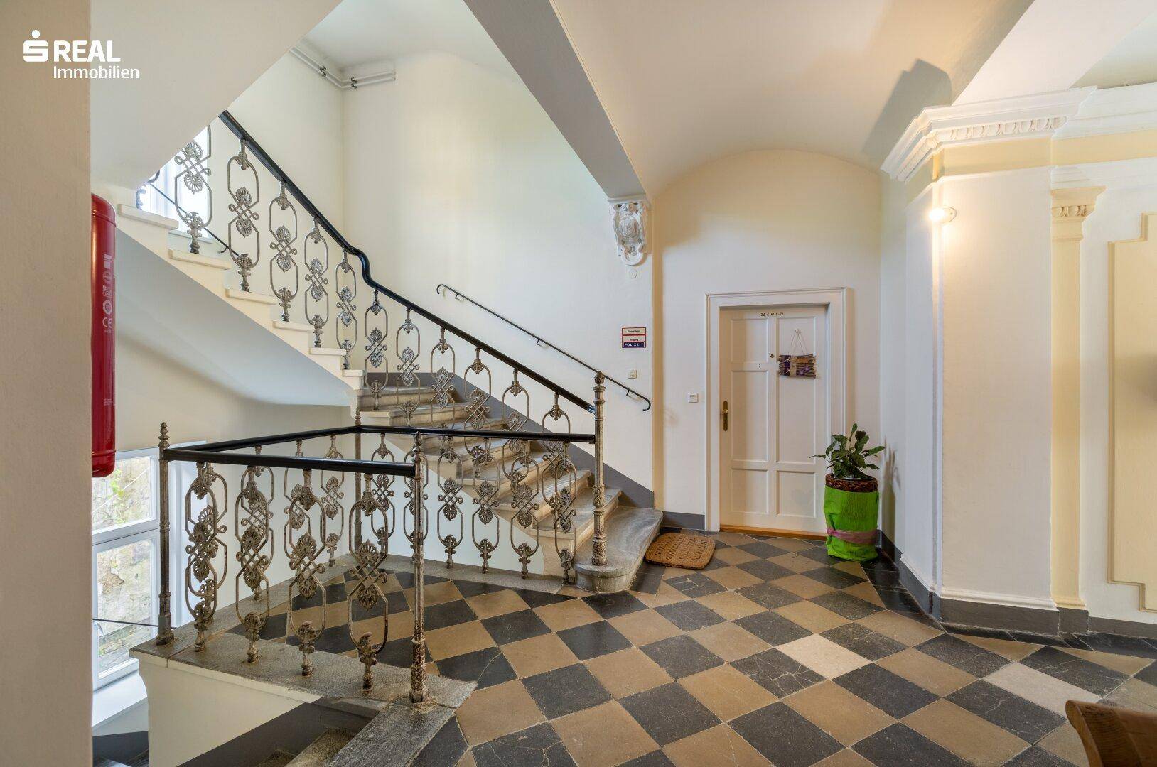 Großzügiges Treppenhaus mit schmiedeeisernem Geländer, hohen Decken und gemustertem Fliesenboden.