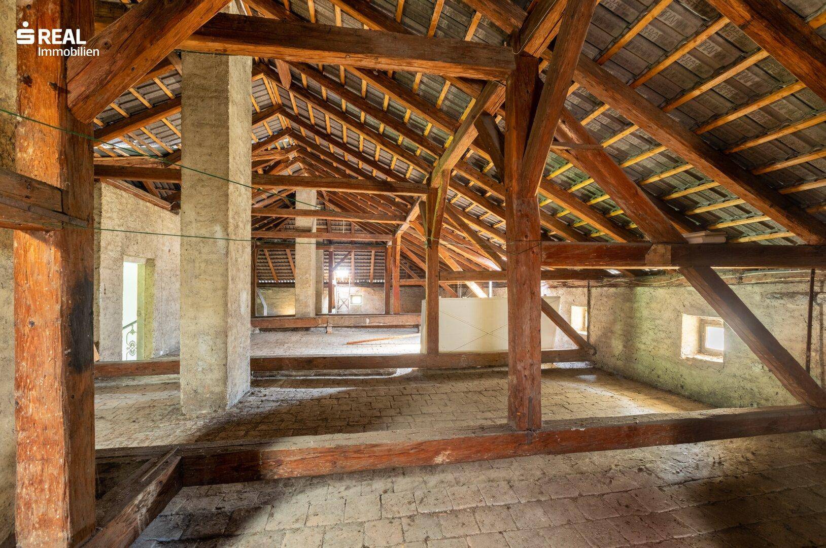 Großer Dachboden mit freiliegenden Holzbalken und natürlichem Lichteinfall durch kleine Fenster.