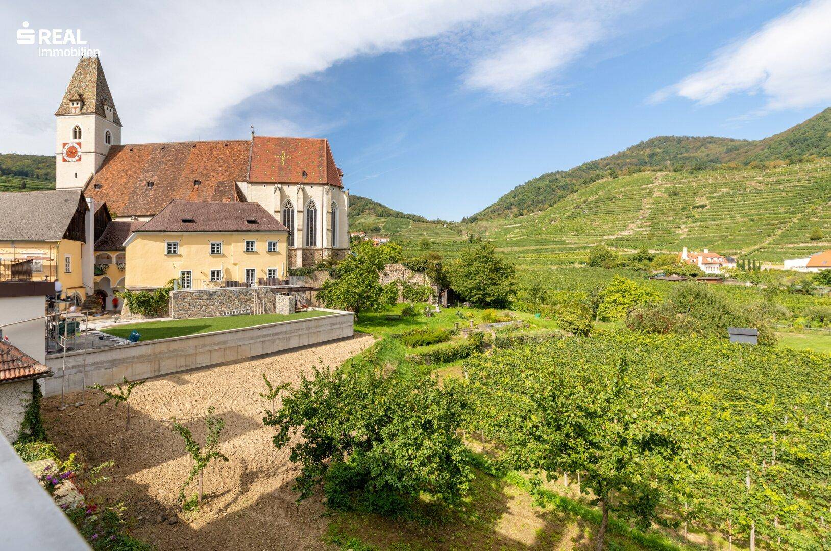 Malerischer Blick auf eine historische Kirche, grüne Weinberge und die umliegende Berglandschaft.