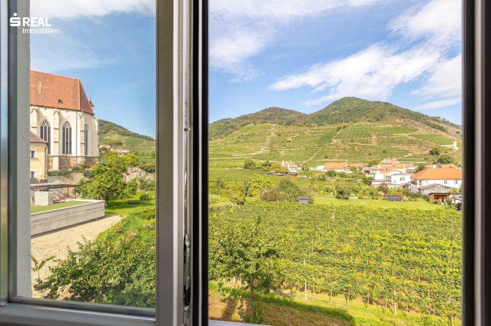 Fensterblick auf eine historische Kirche, weitläufige Weinberge und die beeindruckende Berglandschaft.